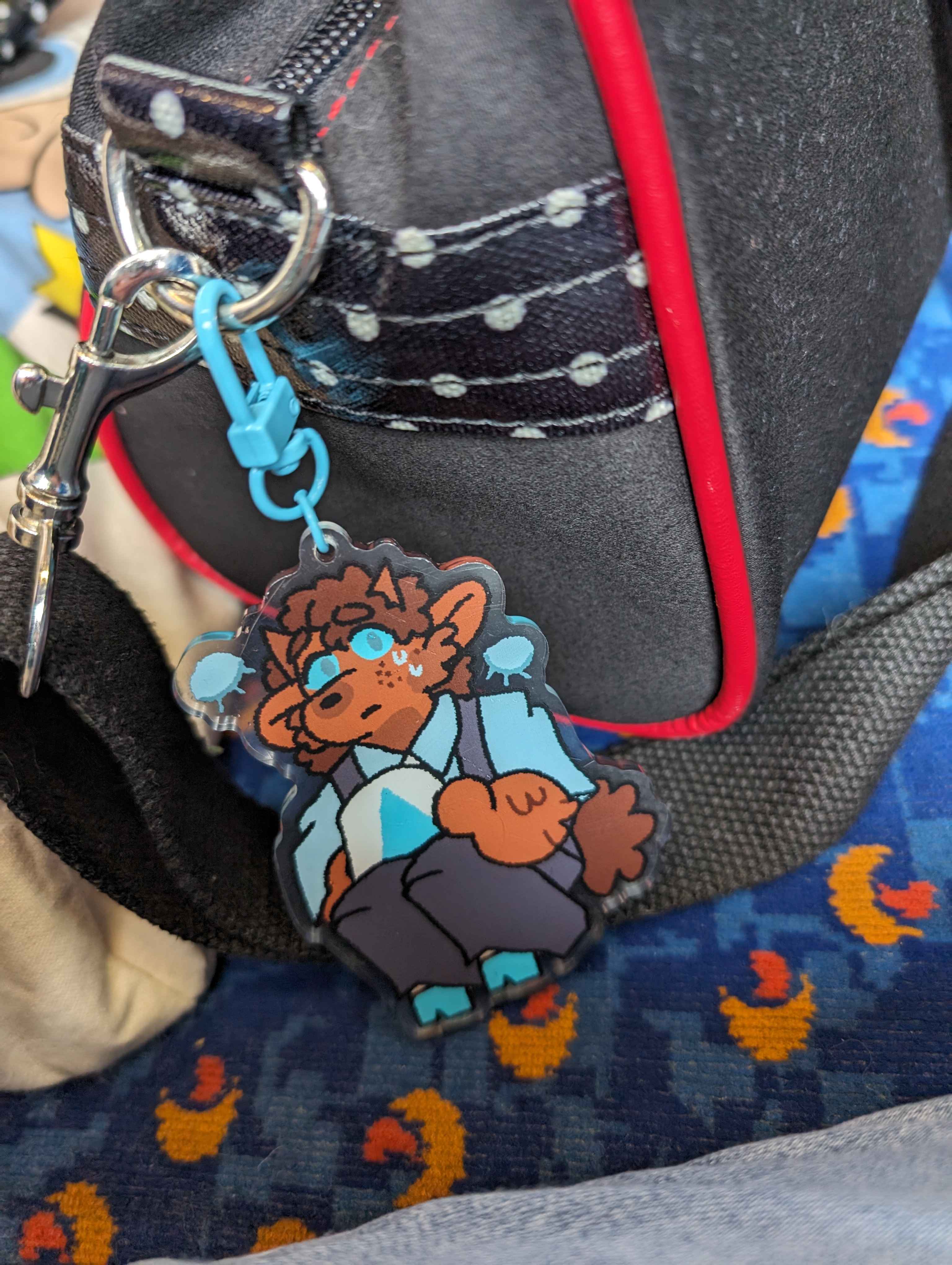 An IRL photo of the Jasper keychain attached to a bag, which is sitting on a bus seat